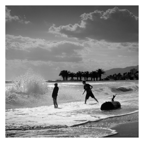 Two boys on a beach, seen backlit by the sun and in silhouette, face a big wave. One stands resolute whilst the other braces himself to run. Washed up on the shore is a big log.