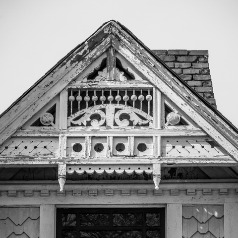 Black and white photograph of an ornate Victorian-era gable end with weathered white painted wood showing extensive peeling and age. The decorative gable features intricate details including turned spindle work in a row near the peak, elaborate scrollwork with two circular medallions on either side, a central carved wooden ornament with curved cutout patterns, four evenly-spaced circular vent holes with vertical grooved panels, scalloped trim work along the lower edge, and decorative brackets supporting the overhang. A brick chimney is visible behind the gable against a plain gray sky. Below the gable is fish-scale shingle siding and a dark window opening.