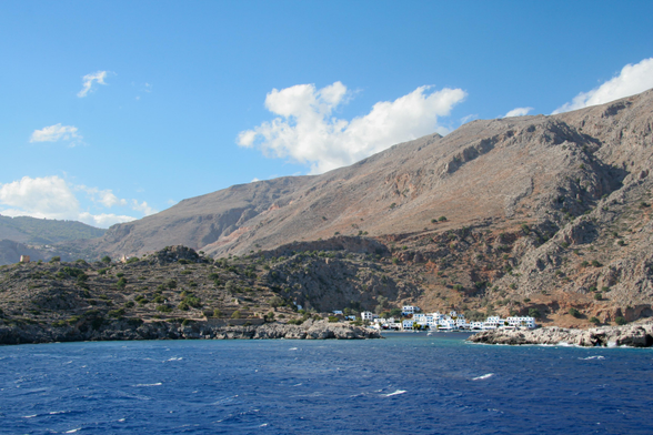 A coastal view featuring a deep blue sea, rugged mountain landscape, and a small cluster of white buildings nestled near the shoreline under a clear, blue sky with scattered clouds.