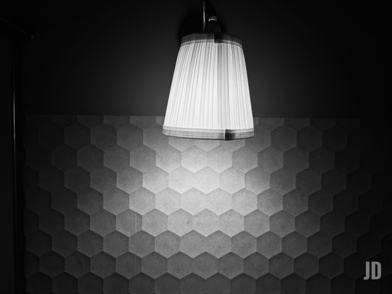 Una lámpara de techo encendida cuelga desde el centro de la habitación, con un diseño moderno y minimalista. Su luz cálida se derrama suavemente sobre una pared decorada con un patrón de hexágonos en diferentes tonos grises, creando un juego de luces y sombras que resalta la textura geométrica. La iluminación genera un ambiente acogedor y elegante, mientras los bordes de los hexágonos parecen tener un leve relieve por el efecto de la luz.