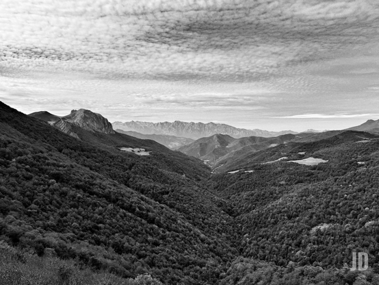 La imagen muestra un paisaje montañoso en blanco y negro, con nubes dispersas que se extienden por el cielo.  En la distancia, se aprecian varios picos, creando una sensación de profundidad y vastedad.  La ausencia de color acentúa las texturas y contrastes de las montañas y el cielo.