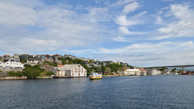A photo of a strait with a small boat going by. There's a hill with houses on it, and a bridge to the right of the photo. The sky is blue with wispy blue clouds.