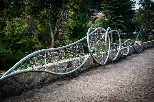 A long, curving metal railing covered in numerous padlocks is the primary subject of the image, set alongside a stone wall and a body of water. The railing, constructed of shiny silver metal, snakes back and forth in a wave-like pattern, densely populated with locks of various sizes and colors. The stone wall beneath the railing appears aged and textured, constructed from irregularly shaped rocks. Beyond the railing and wall is a dark, still body of water, with trees and foliage visible in the background. The ground in the foreground is paved with a light-colored, speckled stone surface, creating a pathway alongside the railing.

Provided by @altbot, generated privately and locally using Gemma3:27b