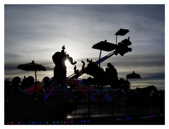 En liggande bild på en åkattraktion på ett nöjesfält. Allt är i silhuett mot himmel, men färgglada lampor syns. Himmelen har slöjmoln.