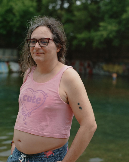 Standing in front of the waters of Barking Springs, Kit O'Connell is wearing a pink crop top with the word "Cute" in a sparkly, dripping heart (from Austin's Cute Nail Studio) and blue swim trunks with red crabs. Their long brown hair is a bit messy, a semicolon tattoo is visible on their left arm and they're wearing red and purple cats eye glasses.  