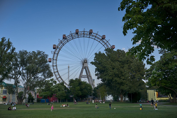 Das Foto zeigt das berühmte Riesenrad im Wiener Prater, einem großen Vergnügungspark in Wien, Österreich. Das Riesenrad ist ein ikonisches Wahrzeichen der Stadt und dominiert die Szene im Hintergrund. Es ist von einem klaren, blauen Himmel umgeben und wird von grünen Bäumen eingerahmt, die dem Bild eine natürliche und entspannte Atmosphäre verleihen.
Im Vordergrund sieht man eine große, grüne Wiese, auf der Menschen entspannt sitzen, spazieren gehen oder sich unterhalten. Einige Kinder spielen auf dem Rasen, was dem Bild eine lebendige und familienfreundliche Stimmung verleiht. Die Szene wirkt friedlich und lädt zum Verweilen ein.