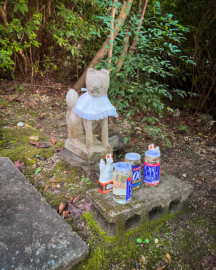 Una pequeña escultura con una figura que, supongo, es un zorro (inari). Junto a la figura, vemos varias latas de sake (las venden en los combini como "one cup sake"). Serán de unos 30cl. Las dejan como ofrenda al dios inari, supongo.