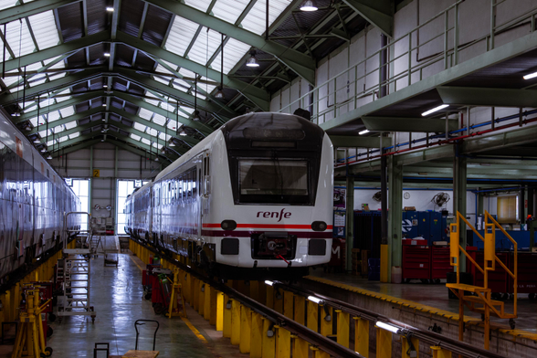 A Renfe class 448 EMU sits on top of some elevated rails, so that workers can inspect it from below, inside a huge industrial warehouse. 