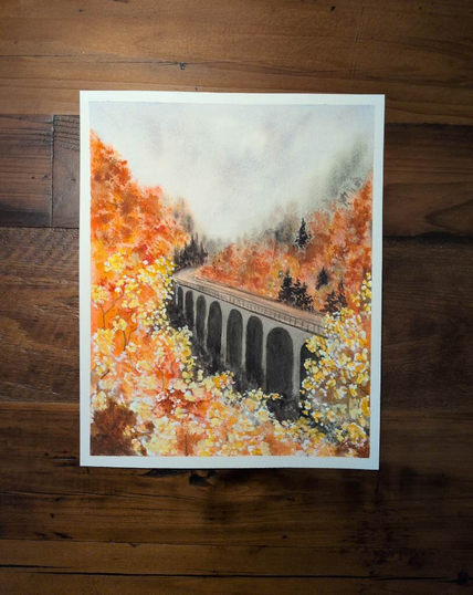 This is an Autumn landscape painting in portrait orientation on a 8x10 sheet of Arches Coldpress watercolor paper. This painting is a November project with Let's Make Art.  The top 1/4 -1/3 is a triangle of textured sky.  Then layers of foliage a little green but mostly oranges and yellows coming from the right bottom is a tall bridge with high arches going at a diagonal to a little more than halfway up and a little left of center where it curves back to the right and disappears. There are also a few evergreen trees in the distance by the roads curve and by the road on the right side. More indistinct evergreens are at the base of the bridge.