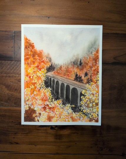 This is an Autumn landscape painting in portrait orientation on a 8x10 sheet of Arches Coldpress watercolor paper. This painting is a November project with Let's Make Art.  The top 1/4 -1/3 is a triangle of textured sky.  Then layers of foliage a little green but mostly oranges and yellows coming from the right bottom is a tall bridge with high arches going at a diagonal to a little more than halfway up and a little left of center where it curves back to the right and disappears. There are also a few evergreen trees in the distance by the roads curve and by the road on the right side. More indistinct evergreens are at the base of the bridge.