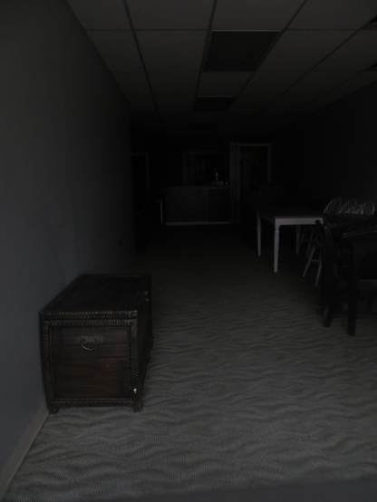A dimly lit room with gray walls and carpet, office tile ceiling, and various chairs, tables, and a chest against the walls. The only light is coming from behind the viewpoint.