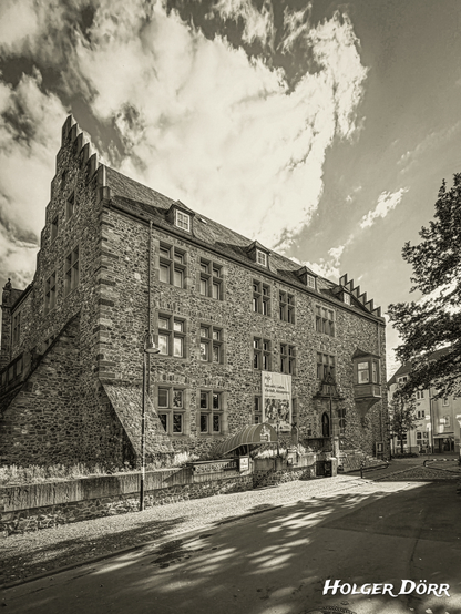 Ein historisches Gebäude aus hellem Bruchstein erhebt sich mächtig unter einem teils wolkigen Himmel. Die Sonne taucht die Fassade des Alten Schlosses in Gießen in warmes Licht und lässt die Struktur des Mauerwerks lebendig wirken. Die steilen Treppen, der spitze Treppengiebel und die markanten Fenster verleihen dem Bau Würde und Zeitlosigkeit. Die Aufnahme in nostalgischem Schwarzweiß lässt die Szene wirken, als stamme sie aus einer anderen Epoche – ruhig, eindrucksvoll und voller Geschichte.