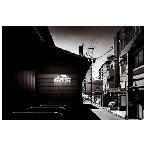 A black-and-white photo of a narrow urban street in Nagoya. Sunlight falls sharply on one side of a wooden building, illuminating a weathered sign with Japanese text while the rest remains in shadow. Power lines crisscross above the street, and apartment buildings line the background. The contrast between bright light and deep shadow gives the scene a cinematic stillness.