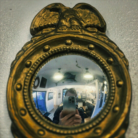 A fisheye view from a convex, nautically themed mirror shows a reflection of me obscured by my iPhone snapping this photograph, the coffee shop bar behind me lined with other nautically themed decor as well.