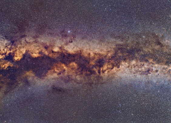 Night time image of a starry sky - many many small stars. Running left to right in the middle of the image is white glow of the Milky Way. There are many dark lanes and dark areas inside the Milky Way (dust obscuring starlight behind). The Milky Way is brighter, wider and more orange'ish on the left.