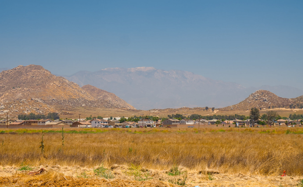 From foreground to background: A field of mostly dry tall grass, many small beige houses, some hills covered by rocks and dry grass, and a great mountain in the haze