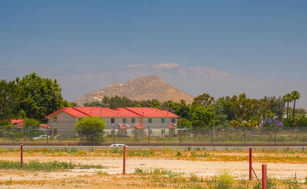 From foreground to background: an empty lot with weeds and some short red poles, a freeway in a ditch with one car popping out, an apartment building with 2 stories and sloped red roof surrounded by trees, a small pointed rocky hill in dry grass, and a great mountain way in the distance in the haze