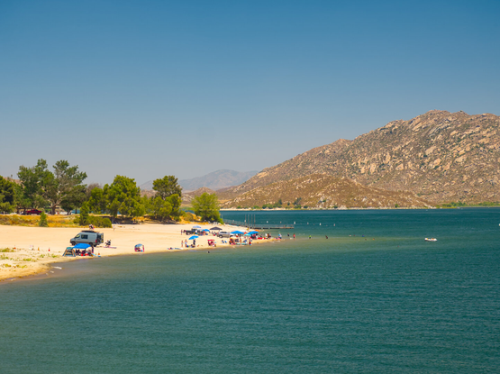 A beach and trees are by a vast artificial lake. Many people were playing on the beach and shallow parts of the lake. An arid hill rises above the lake in the distance.