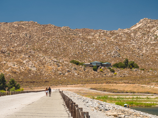 A couple were walking on the long dam top of Lake Perris. The dam top leads to a backdrop of an arid mountain. A modern building (Ya'i Heki' Regional Indian Museum) perches on the hill.