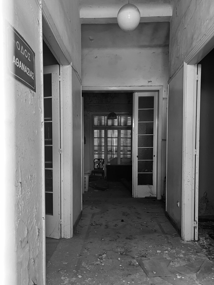 Black and white photo. The interior of an abandoned house, taken through a gap in the locked door. Front hall, looking through to open glass doors and a large shuttered window. All plain and painted white. To left and right open doorways. A white globe light hangs above. On the wall to the left a street sign reads Immortality Street in Greek.