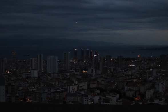 Eine Nachtansicht einer Stadt mit beleuchteten Gebäuden und einer Küstenregion im Hintergrund. Der Himmel ist bewölkt, und auf dem Wasser sind mehrere Boote zu sehen.