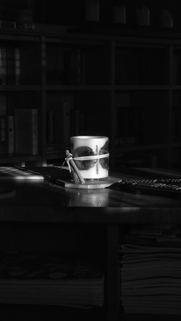 Schwarzweißfoto. Im Zentrum des Bildes steht eine dicke Stumpenkerze auf einer Tischplatte in einem Raum. Sie ist mit einem Bastband umwickelt, in das auf der linken Seite eine Zimtstange eingebunden ist. Die Kerze brennt nicht, sondern wird von einfallendem Licht angestrahlt. Der Rest des Raumes und des Inventars liegt im Dunkeln und ist nicht oder nur schwer zu erkennen.