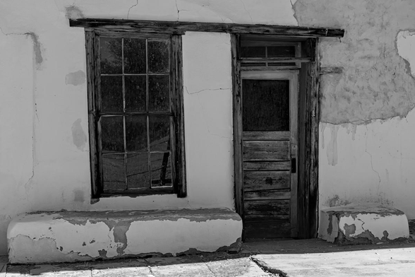 A black and white landscape photo of a weathered wood framed window, on the left, and door on the right. The header beam cross from the window to the door. The window and door are set in the white adobe wall that has large parts of the plaster missing in the upper right. The wall is in shade and there's a beam of sunlight across the sidewalk in front of the wall with the window and door.