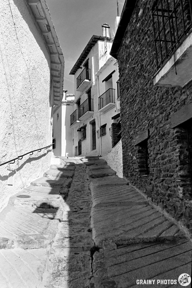 Black and white photo of a narrow, steep cobbled street in Capileira, lined with white and stone buildings. Windows have bars and balconies. Shadows create dramatic contrast.
