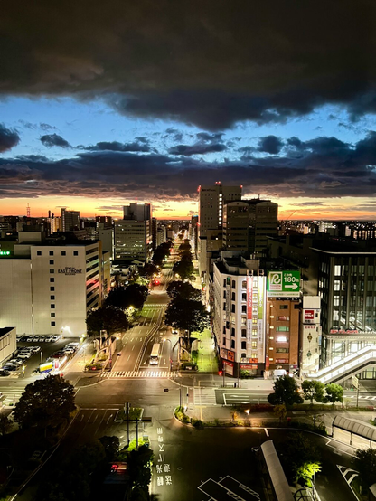 A dramatic orange sunrise under dark clouds and just above high-rise office buildings along a wide, tree-lined city street.