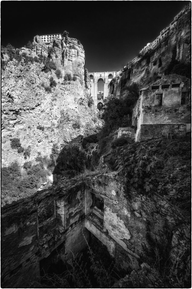 Ein tiefes, steiles Tal mit einer imposanten Brücke, die sich hoch über dem Abgrund spannt. Links ragt ein Felsen mit einem Gebäude darauf empor. Rechts und im Vordergrund führen alte, verwitterte Mauern und Ruinen den Hang hinunter. Die Szene wirkt dramatisch durch das Spiel von Licht und Schatten.
