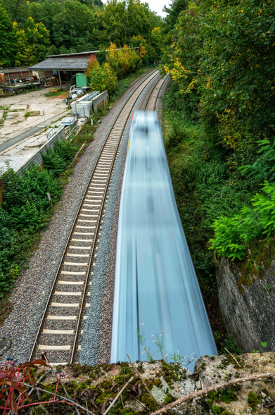 Auf dem Bild ist eine Eisenbahnstrecke zu sehen, auf der ein Zug mit hoher Geschwindigkeit fährt. Der Zug ist aufgrund der Bewegung verschwommen dargestellt. Die Perspektive ist von oben, und man blickt auf die Gleise, die von Bäumen und Sträuchern umgeben sind. Am linken Bildrand befindet sich ein Gebäude mit einem Schotterplatz.