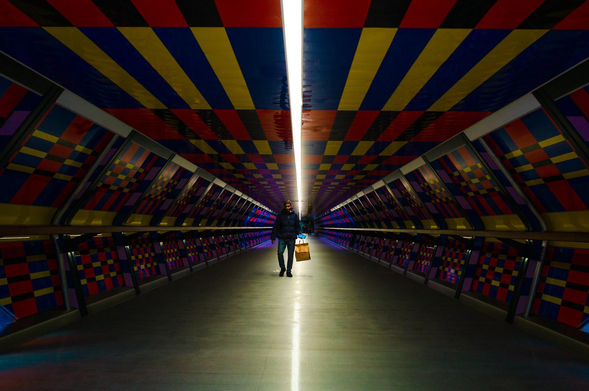 The image captures a person walking down a vibrantly decorated tunnel. The tunnel's most striking feature is its ceiling and walls, which are adorned with a repeating pattern of colorful squares and stripes in shades of red, blue, yellow, and black. A bright, linear light source runs down the center of the ceiling, illuminating the path and creating a strong sense of perspective.

The person is the central subject, positioned in the middle of the tunnel, walking towards the viewer. They are wearing a dark jacket, jeans, and black shoes. They are carrying a shopping bag.

The tunnel's design leads the eye towards the vanishing point, creating a strong sense of depth. The walls have a railing running along them, and are lined with glass panels. The floor is a flat, neutral grey.