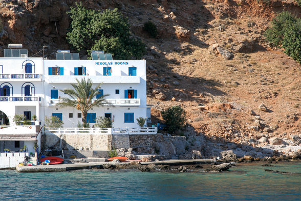 White building labeled “Nikolas Rooms” by the waterfront, with blue shutters and doors, against a rocky hillside and clear blue water in the foreground.