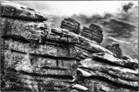 Schwarz-Weiss-Fotografie schroffer, geschichteter Felsformationen im Vordergrund, die durch Erosion geformt wurden. Im Hintergrund eine leicht hügelige Landschaft, die sich in sanften Wellen bis zum Horizont erstreckt. Wolken hängen tief über den Hügeln und verleihen der Szene eine mystische Stimmung.