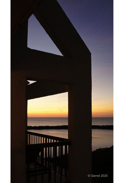 Silhouette of a balcony railing against a vibrant sunset with purple and orange hues over a calm ocean seascape, conveying tranquility.