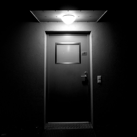 Das Foto zeigt eine metallene Haustür. Die Tür ist mit einem kleinen, undurchsichtigen Fenster ausgestattet und wird von einer zentralen Leuchte eines Vordaches direkt angestrahlt. Der Türgriff und das Schloss sind funktional gestaltet. Der Boden vor der Tür besteht aus einem Metallgitter, während die Wände glatt und ohne Dekoration sind. Die Schwarz-Weiß-Aufnahme betont den Kontrast zwischen dem beleuchteten Türbereich und der vorherschenden Dunkelheit. Die Atmosphäre wirkt sachlich und karg.