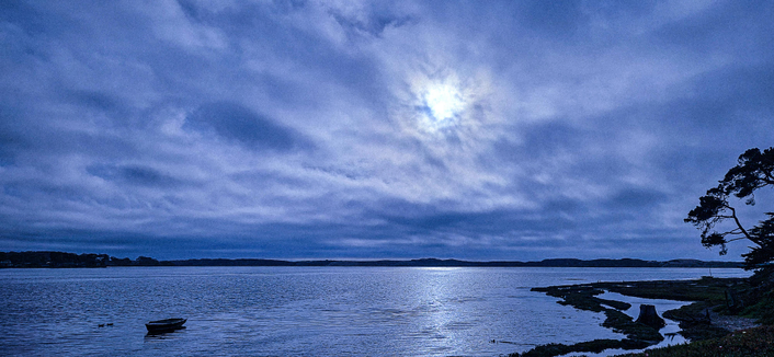  A wide, panoramic view of a body of water under a cloudy, dark blue sky. The water stretches across the majority of the frame, reflecting a bright, glowing spot in the sky, likely the moon or sun obscured by clouds. A dark shoreline with rocks and sparse vegetation runs along the right side of the image, extending into the distance. A small, dark boat is visible on the left side of the water, near the horizon. The land in the background is a dark, indistinct shape against the bright sky.