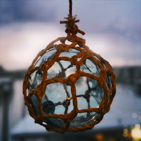 A glass sphere wrapped with rope in decorative knots hangs against a window, backlit by an overcast sky.