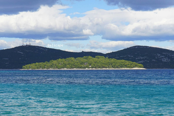 Fotografia przedstawiająca jedną z wysp w pobliżu wyspy Murter. Została zrobiona właśnie z tej wyspy. Za tą wyspą może być archipelag Kornat lub pasmo górskie kontynentu Dalmacji, nie wiadomo. Wyspa znajduje się w centrum zdjęcia. Kolor morza pionowo pod wyspą jest w większości turkusowy. Morze jest spokojne chociaż drobne fale przez lokalne podmuchy są widoczne. Wyspa w większości jest porośnięta zieloną roślinnością. U jej podnóża widoczne jest niewysokie pasmo skalistego brzegu koloru kremowego. Niebo nad wyspą jest w połowie pochmurne, ale jest bardzo jasne.