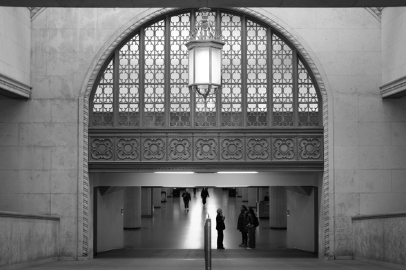 Looking down the ramp to the curved arch leading to the VIA Rail concourse.  Above is an intricately designed fan light surrounded by carved patterns in the stone. In the opening are framed various customers and employees, small in the distance. Down the centre of the concourse are two bright lines of reflected light, leading into the distance.
