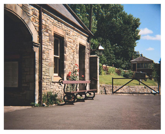 Vintage looking colour film photograph showing an empty brown bench outside a railway station waiting room.  