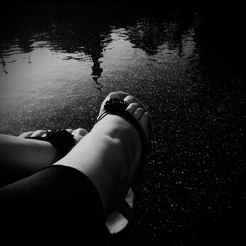 Ein Schwarzweißfoto zeigt ein Paar Füße in Sandalen, die am Rand eines Gewässers ruhen. Im Wasser spiegelt sich unscharf einer städtischen Bebauung, während Lichtreflexe auf der Oberfläche tanzen.