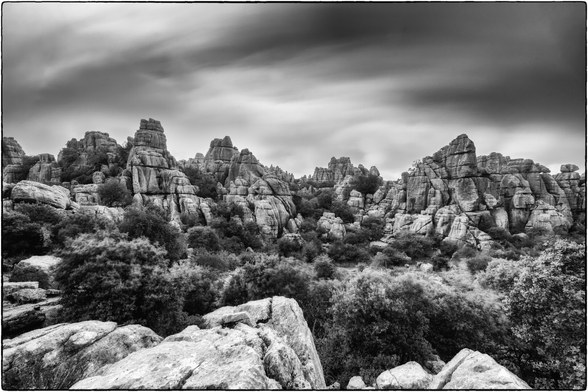 Schwarz-Weiss-Aufnahme einer weiten, zerklüfteten Karstlandschaft, aufgenommen in einer Langzeitbelichtung, die eine dramatische Atmosphäre erzeugt.

Im Vordergrund liegen grosse, helle Kalksteinbrocken, deren Textur und Bruchstellen detailreich hervorgehoben sind. Dahinter erstreckt sich eine hügelige Ebene, die von dichtem, struppigem Gebüsch und kleineren Bäumen bedeckt ist.

Der Mittel- und Hintergrund wird von einer langen Reihe bizarrer, gestapelter Felsformationen dominiert. Diese Felsen sind horizontal geschichtet und wirken wie eine natürliche Festung.

Der Himmel ist durch die Langzeitbelichtung verschwommen und streifig, was die Bewegung der Wolken andeutet. Die dunklen, dramatisch gezeichneten Wolken bilden einen starken Kontrast zu den hellen Felsen und verleihen der gesamten Szene eine epische und zugleich düstere Stimmung.