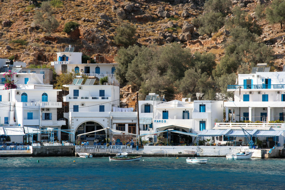 Seaside village with white buildings and blue shutters nestled against a rocky hillside. Small boats are moored in the clear blue water.