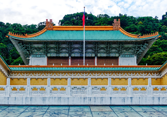 Palace side building with a wall and a flagpole in front, trees in the background.