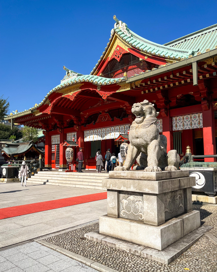 Vista diagonal de la fachada del santuario principal (del estilo habitual: madera lacada en rojo, techos curvados de tejas verdes y adornos dorados, etc). En primer plano se ve una pequeña escultura de piedra de lo que podría ser un dragón. Hay una alfombra roja que lleva a la entrada del templo (creo que había alguna celebración en marcha).