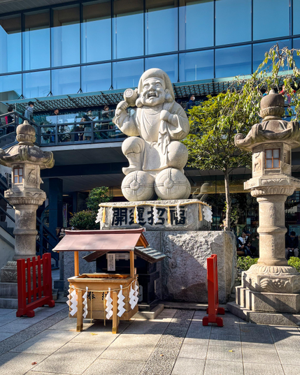 Una escultura de piedra blanca que, supongo, representa algún dios (Daikokuten, creo). A mí me recuerda a uno de los enanos de Blancanieves: con barba, un saco a la espalda y un mazo en la mano :) A los lados hay un par de linternas de piedra (de adorno, supongo).