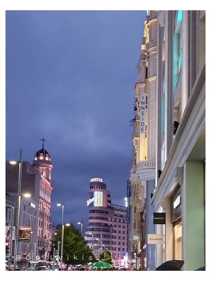 Vista del letrero de Schweppes desde la lejanía (mitad negro y mitad de colores). Hay altos edificios a ambos lados. El cielo está plomizo por la hora y las nubes cargadas de lluvia. Las luces de los comercios destacan por su luminosidad y las farolas ya están encendidas.