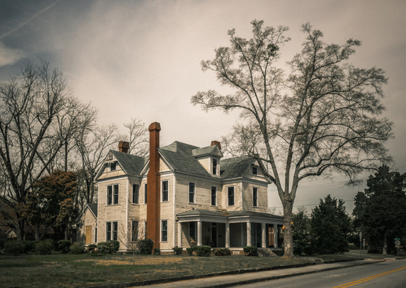Weathered two-story Victorian house with cream-colored horizontal siding and multiple steep gabled roofs under a dramatic overcast sky. The home features a wraparound front porch, tall narrow windows with dark frames, and three prominent brick chimneys in varying shades of red and brown. Bare deciduous trees frame both sides of the structure, with mature oaks showing intricate branch patterns against the moody sky. The front yard shows patchy brown grass with low evergreen shrubs planted along the foundation. A paved road with yellow center lines runs along the foreground, and a boarded-up window is visible on the left side of the house, suggesting vacancy or disrepair.
