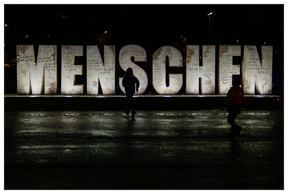 Vor hell leuchtenden Buchstaben, die das Wort "MENSCHEN" buchstabieren, laufen zwei als Silhouetten abgebildete Nachwuchsmenschen. 